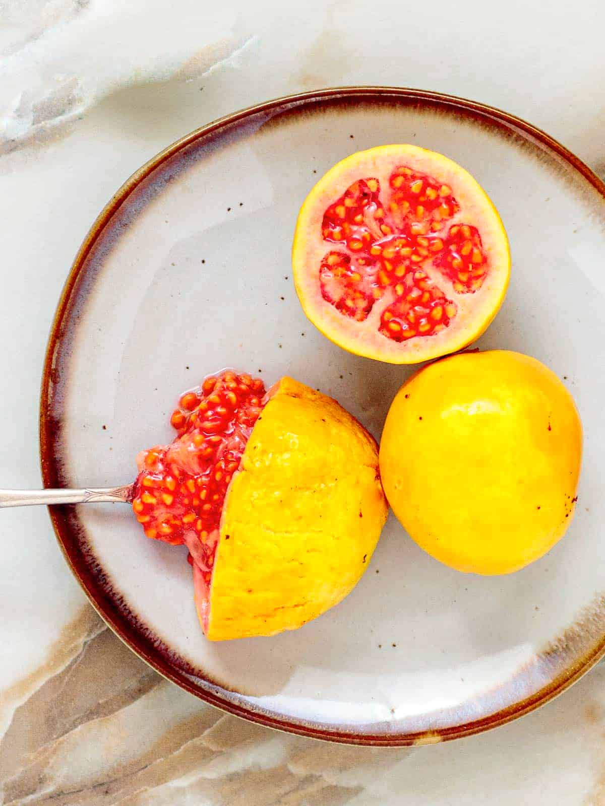 Two guavas on a plate with one showing juicy ripe pink flesh on a spoon.