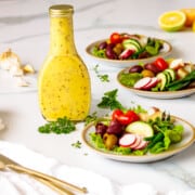 Bottle of homemade lemon Dijon dressing with herbs next to salad plates.